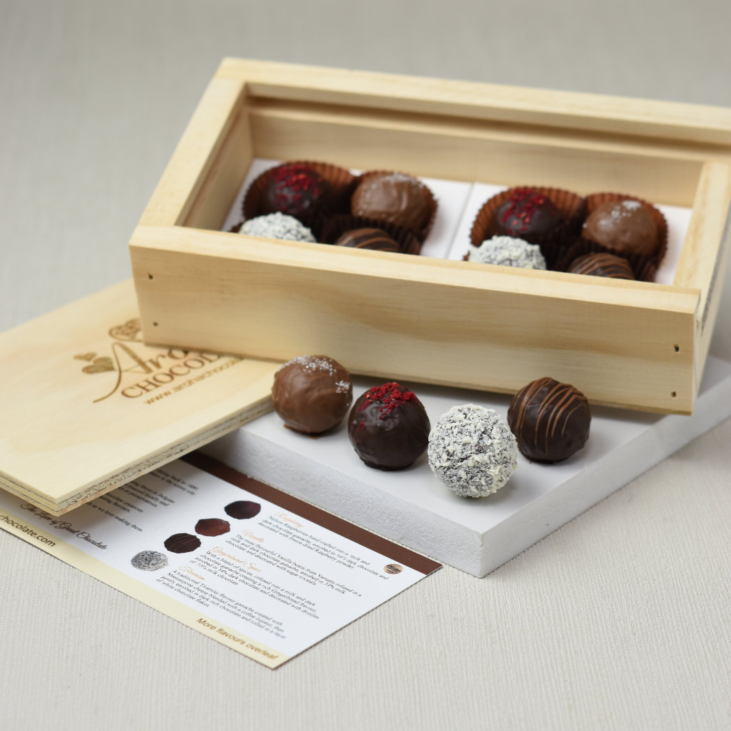 Aroha Chocolate Eight Mixed Truffles Luxury Wooden Gift Box. An open wooden gift box containing beautiful hand crafted chocolates, with the sliding lid sitting below the box. The included flavour chart is beside the box. One of each truffle flavour is out on display in front of the box.