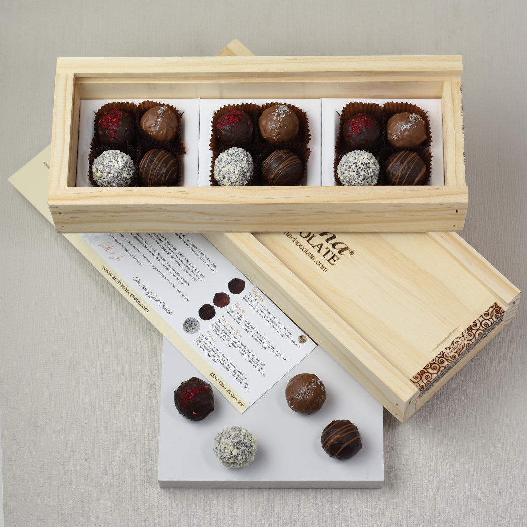 Aroha Chocolate Twelve Mixed Truffles Luxury Wooden Gift Box. Two boxes stacked on top of each other, the top one is open showing the beautiful hand made chocolates, while the bottom box is still closed. There is a menu card and one of each of the chocolate truffles on display in front of the gift boxes.